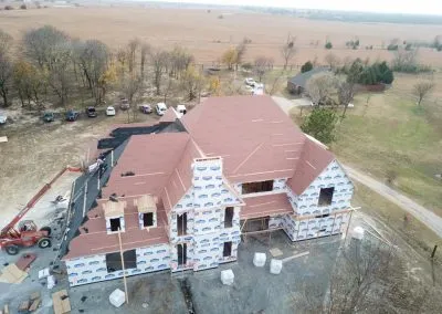 A bird's-eye view of a partially constructed house in a rural area. The house's structure is mostly complete, with a roofing contractor visibly at work. Construction materials and vehicles are nearby, and the surroundings include open fields and scattered trees.