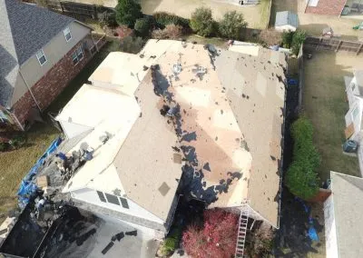 Aerial view of a residential house undergoing roof replacement by a roofing contractor. The old shingles are partially removed, revealing the wooden base underneath. Shingle debris is scattered around the house, with workers present on different sections of the roof and ladders around the house.