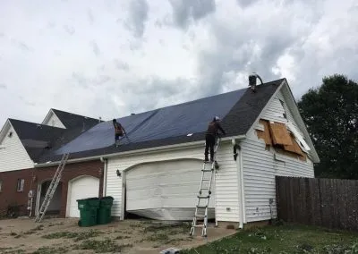 Three men from a roofing contractor are seen repairing a partially damaged roof of a house. Two are on ladders while another is on the roof, securing a large black tarp over the damaged area. The garage door is also damaged, and there are green trash bins by the side of the house.