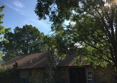 A person is sitting on the roof of a house with stone walls, beneath a canopy of trees. A chimney juts out from the roof, and a ladder is positioned against the wall of the house. Bathed in sunlight with a clear blue sky above, this could be a serene project for any roofing company Tulsa trusts.