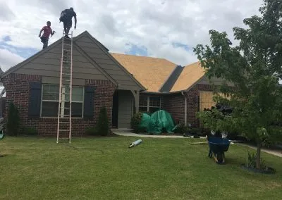 Two workers from a roofing company Tulsa are on the roof of a brick single-story house, one climbing a ladder. The roof is partially covered in plywood, indicating ongoing repairs. The yard has tools, materials, and a wheelbarrow on the grass. Bushes and a tree frame the scene under a cloudy sky.