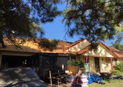 A house with a red trim is having its roof repaired by a roofing contractor. One worker is visible on the roof, while another stands next to a black dumpster in the driveway. Various roofing materials are scattered around, including rolls of underlayment and shingles. Trees frame the scene.