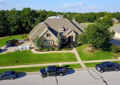 A large, two-story stone and brick house with a neatly manicured lawn and several trees. Multiple vehicles, including trucks with Roofing Tulsa company logos, are parked in the driveway and on the street. Roofing materials are visible on the lawn and roof.
