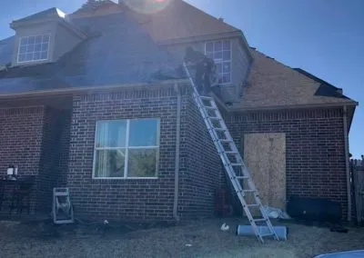 Two workers from a roofing company Tulsa are on a ladder and roof of a brick house, repairing the roof. The sun is shining brightly from behind the house, creating a lens flare. There's a blue tarp on the grass and various tools and materials are scattered around.